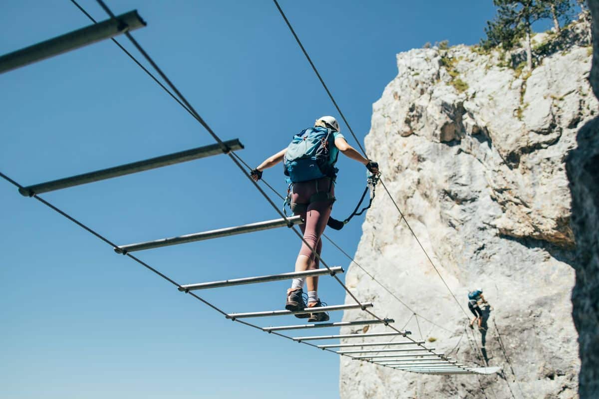 Mujer cruzando un puente colgante en una vía ferrata, equipada con arnés y casco, con un fondo de pared rocosa.