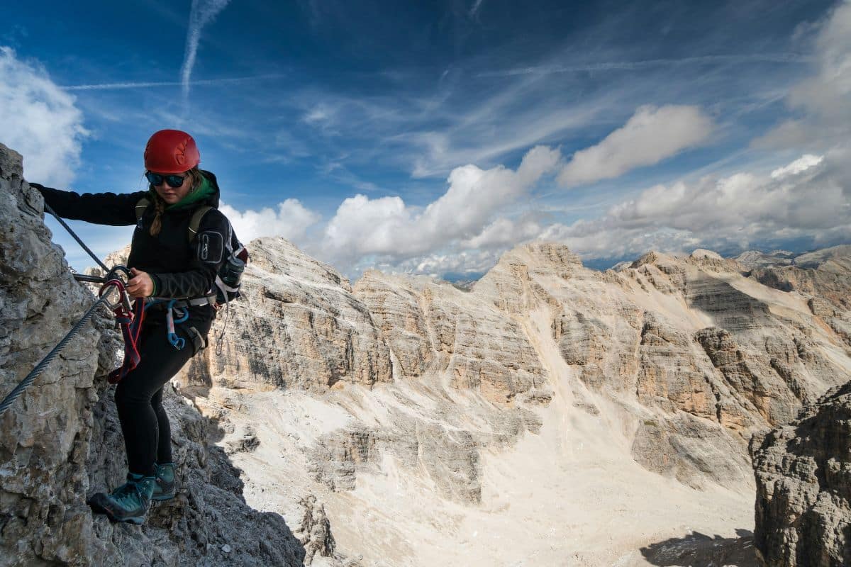 Mujer con casco rojo y arnés asegurada a un cable en una vía ferrata con montañas rocosas de fondo.