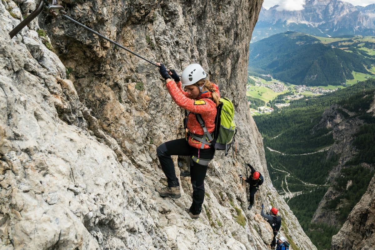 Mujer con chaqueta roja y casco blanco subiendo por una vía ferrata, asegurada a un cable, con otros escaladores detrás y un valle verde al fondo."