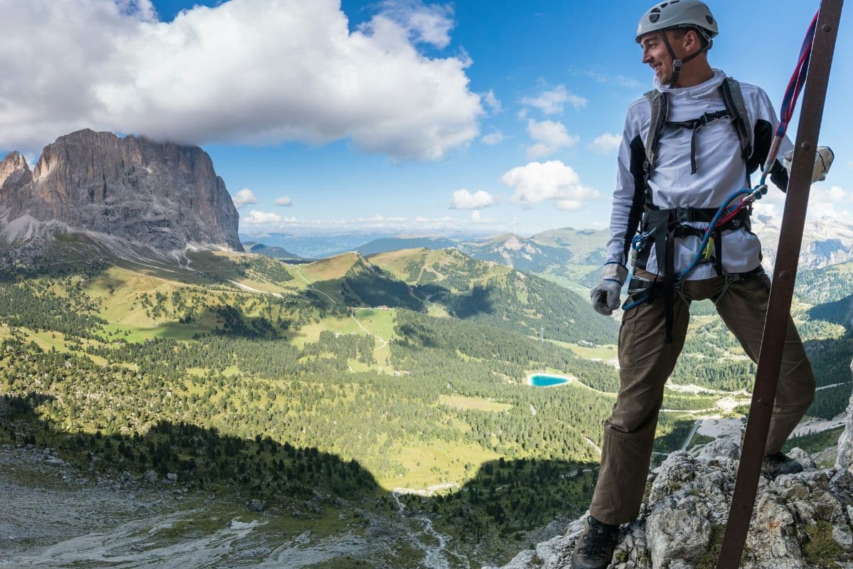 Hombre con casco y arnés asegurado a un cable en una vía ferrata en las Dolomitas, con un impresionante paisaje alpino y un lago azul al fondo.