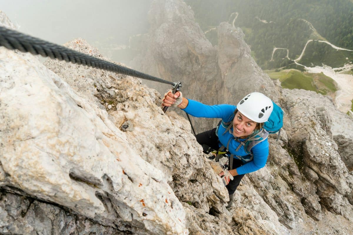 Mujer con chaqueta azul y casco blanco ascendiendo por una vía ferrata, asegurada al cable de acero, con un paisaje de montañas y senderos al fondo.